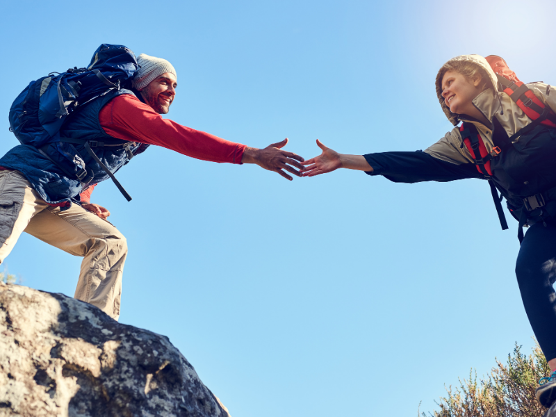 Two hikers reaching out to help each other climb a rocky ledge, representing perseverance and teamwork.